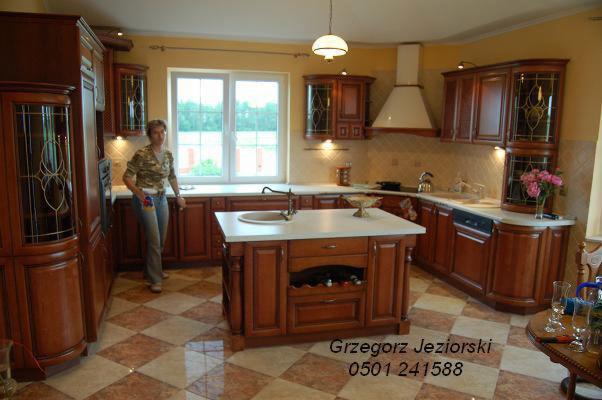 Klasyczna kuchnia z drewnianymi szafkami, wyspą z białym blatem i widoczną osobą w tle, na podłodze wzór szachownicy.