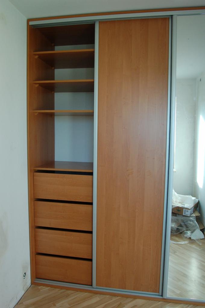 Jasna, drewnopodobna szafa przesuwna z lustrem i otwartą częścią z półkami i szufladami, widoczna na tle pokoju w trakcie remontu.
