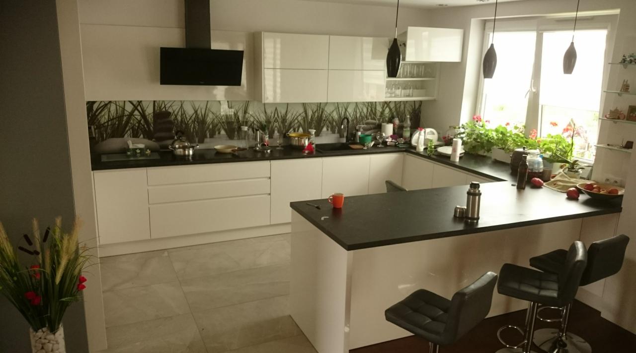 Nowoczesna biała kuchnia z czarnym blatem i wyspą, ozdobiona grafiką roślinną na ścianie, oświetlona wiszącymi lampami i naturalnym światłem z okna, z dodatkami w postaci ziół i owoców na blacie.