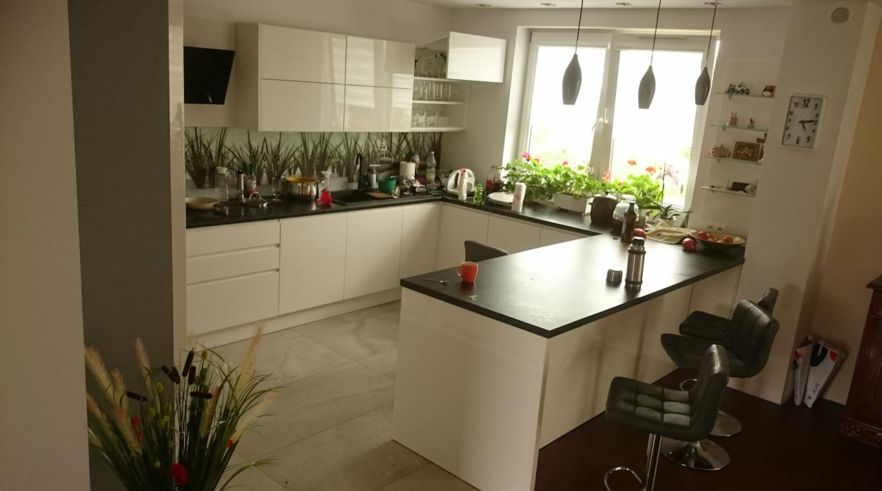 Jasna kuchnia w stylu nowoczesnym z białymi frontami, czarnym blatem, wyspą i hokerami, z oknem z widokiem na zieleń i dekoracyjnym panelem szklanym z motywem roślinnym nad blatem.