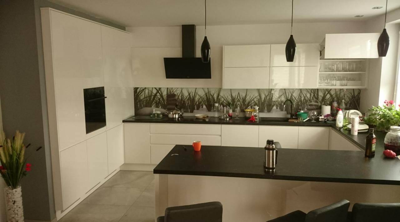 Nowoczesna, biała kuchnia z czarnym blatem i wyspą, oświetlona wiszącymi lampami. Na blacie widoczne naczynia, przyprawy i roślina doniczkowa. Ściana nad blatem wykończona panelem z motywem trawy.