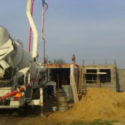 Betonomieszarka z pompą podaje beton na budowę domu z pustaków, widoczni robotnicy i sterta piasku.