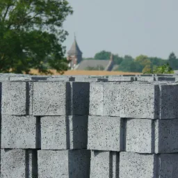 Ułożone w stosy szare bloczki betonowe o porowatej strukturze, w tle drzewa i zarys kościoła.