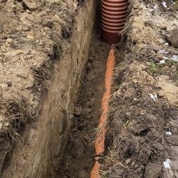 HYDROMAX Grażyna Kotynia - Pomarańczowa rura kanalizacyjna ułożona w wykopie ziemnym, widoczny pracownik w pomarańczowej kurtce. Częściowo zasypana ziemią, w tle resztki śniegu.
