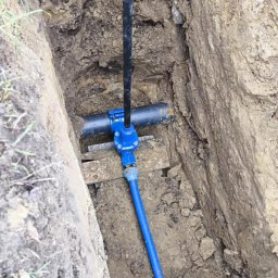 HYDROMAX Grażyna Kotynia - Wykop z widoczną instalacją wodociągową z niebieską rurą i czarną pionową rurą zakończoną elementem z tworzywa sztucznego, stabilizowaną drewnianymi rozpórkami.