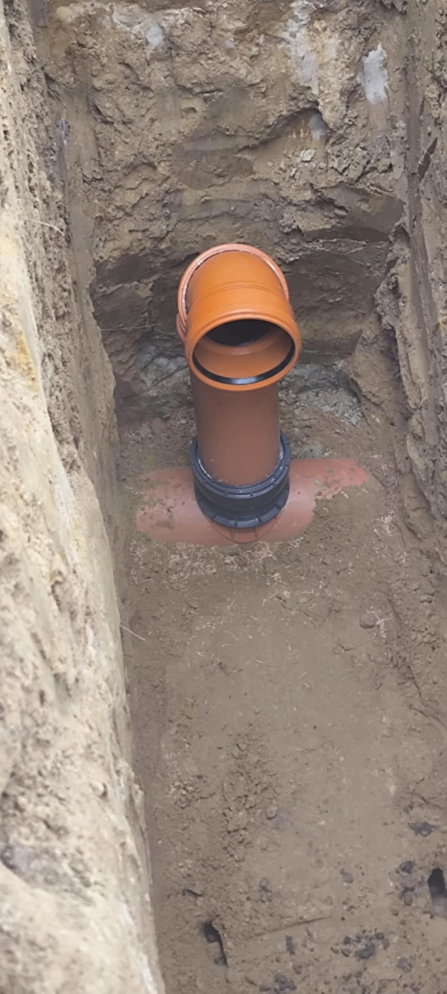 Pomarańczowa rura kanalizacyjna z kolankiem, połączona z poziomą rurą, umieszczona w wykopie ziemnym o pionowych ścianach.