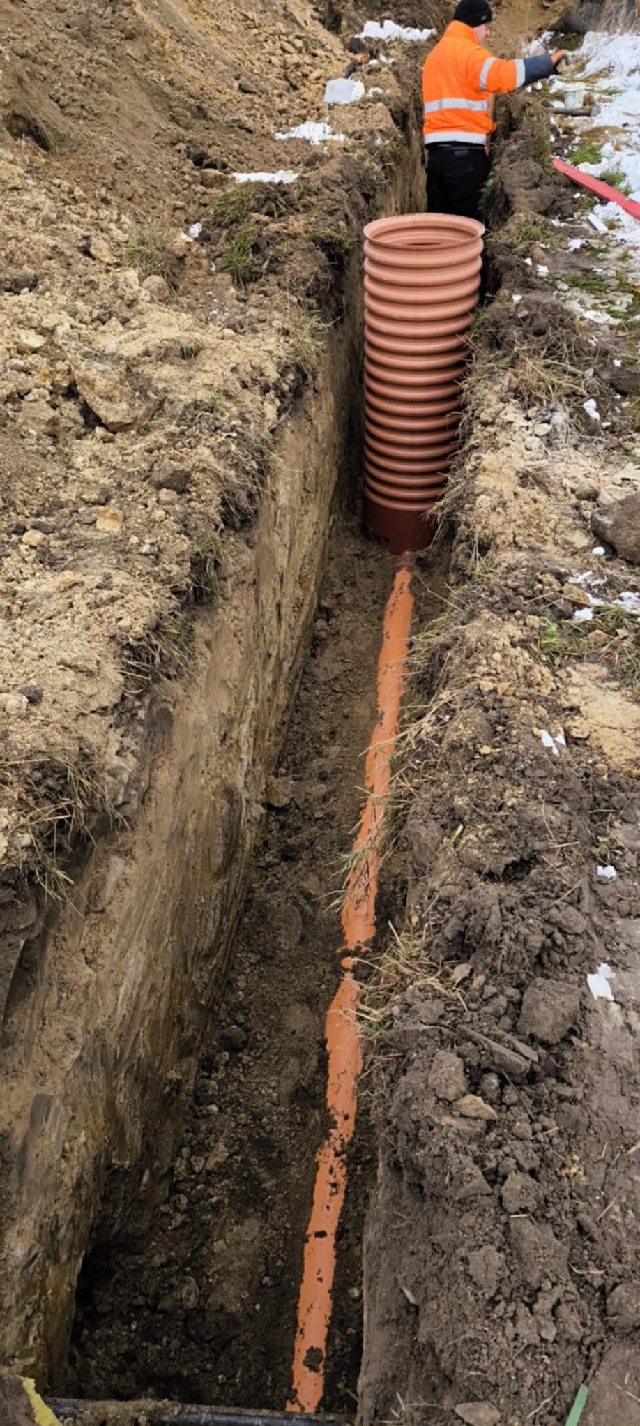 Pomarańczowa rura kanalizacyjna ułożona w wykopie ziemnym, widoczny pracownik w pomarańczowej kurtce. Częściowo zasypana ziemią, w tle resztki śniegu.