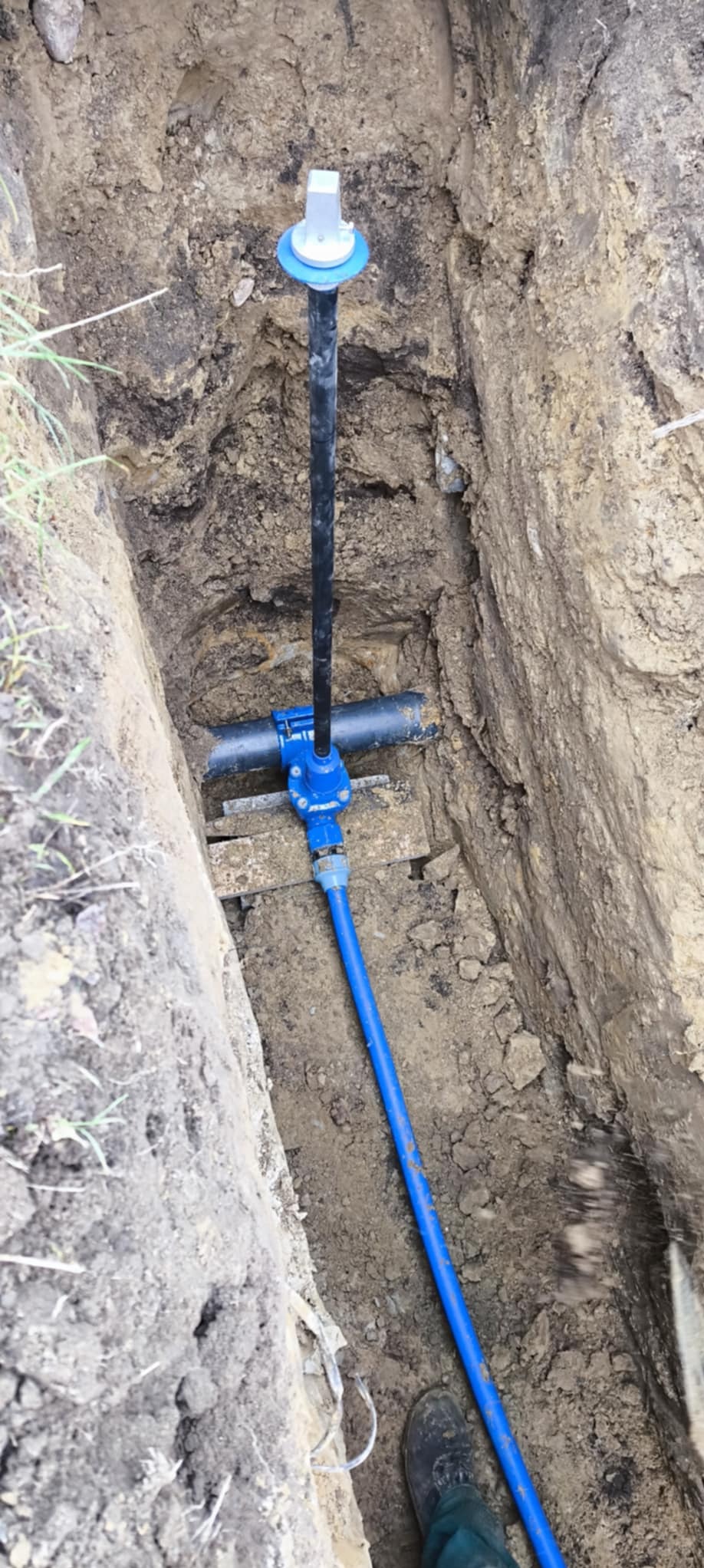 Wykop z widoczną instalacją wodociągową z niebieską rurą i czarną pionową rurą zakończoną elementem z tworzywa sztucznego, stabilizowaną drewnianymi rozpórkami.