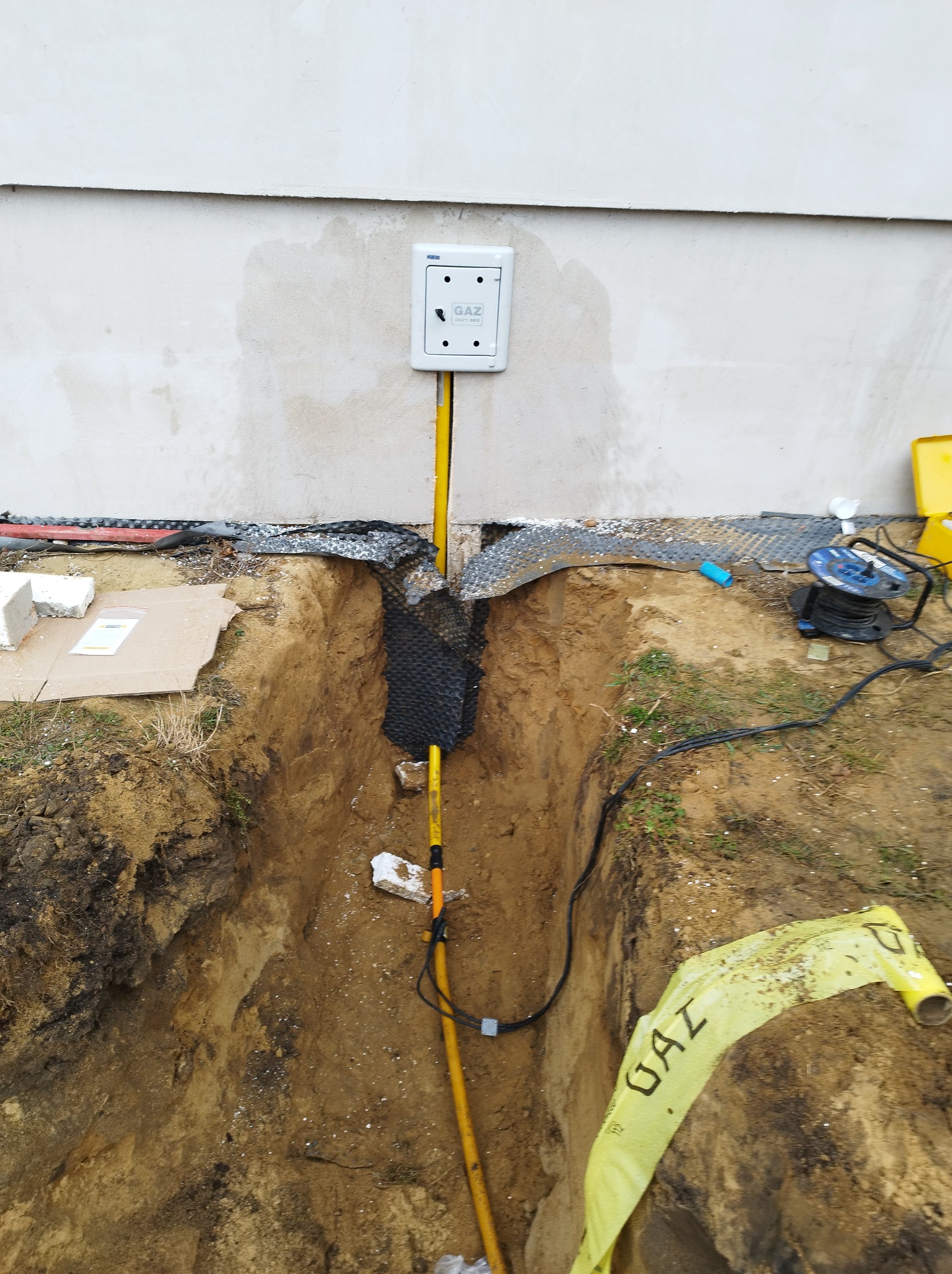 Wykonany wykop pod przyłącze gazowe do budynku, widoczna żółta rura gazowa w ziemi oraz skrzynka przyłączeniowa na ścianie budynku.