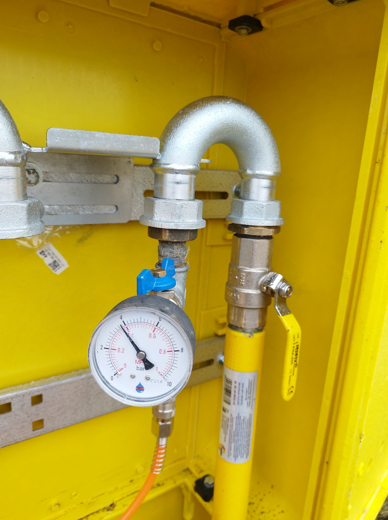 Żółta skrzynka gazowa z widocznymi elementami instalacji: srebrne rury, zawór z żółtą rączką, manometr z niebieskim zaworem oraz pomarańczowy przewód.