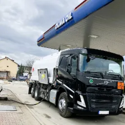 Czarna cysterna Volvo pod dystrybutorem stacji paliw Olma Oil, z podłączonym wężem do podziemnego zbiornika.