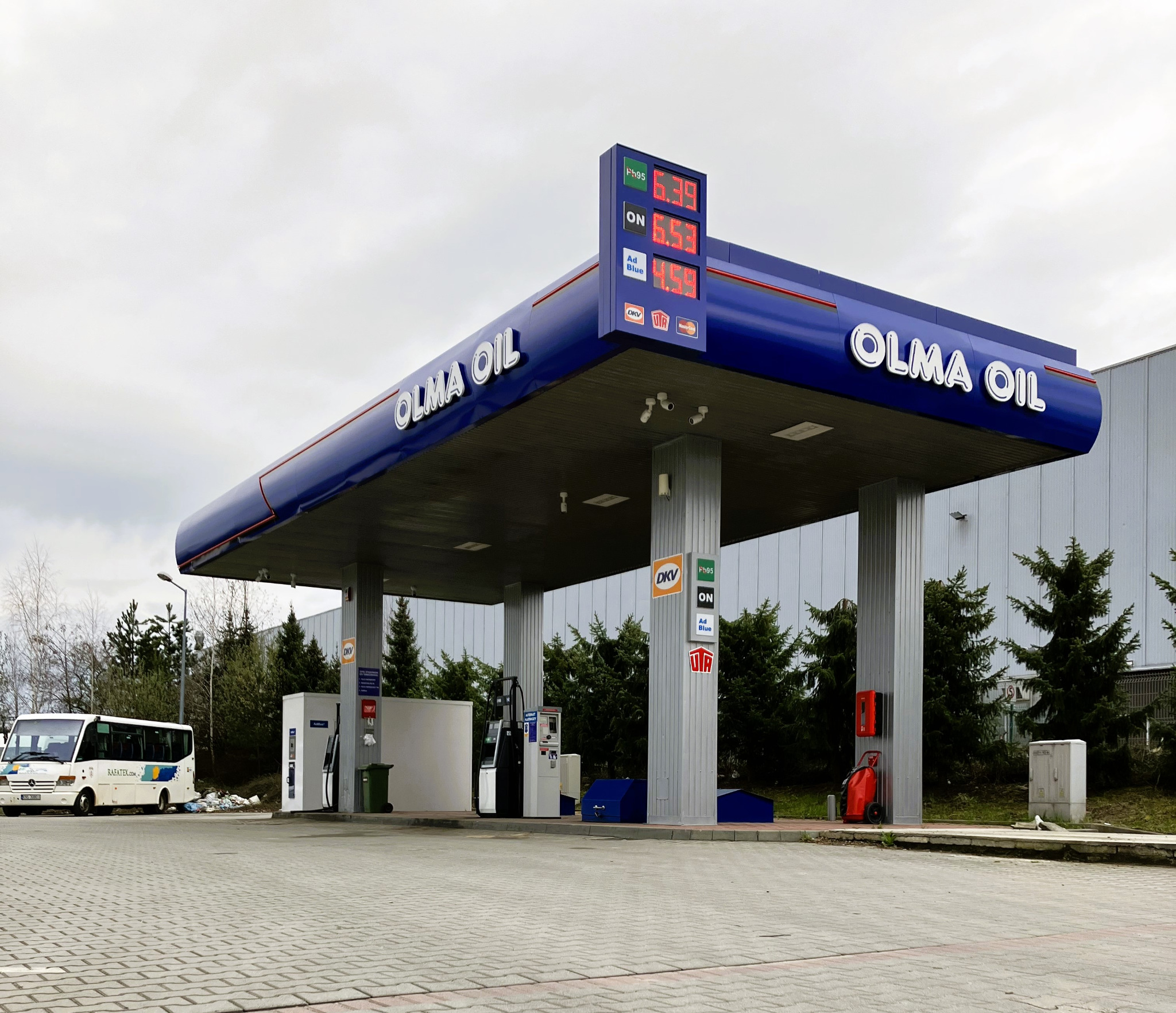 Stacja paliw Olma Oil z widocznymi dystrybutorami, cennikiem i autobusem w tle, sfotografowana w pochmurny dzień.