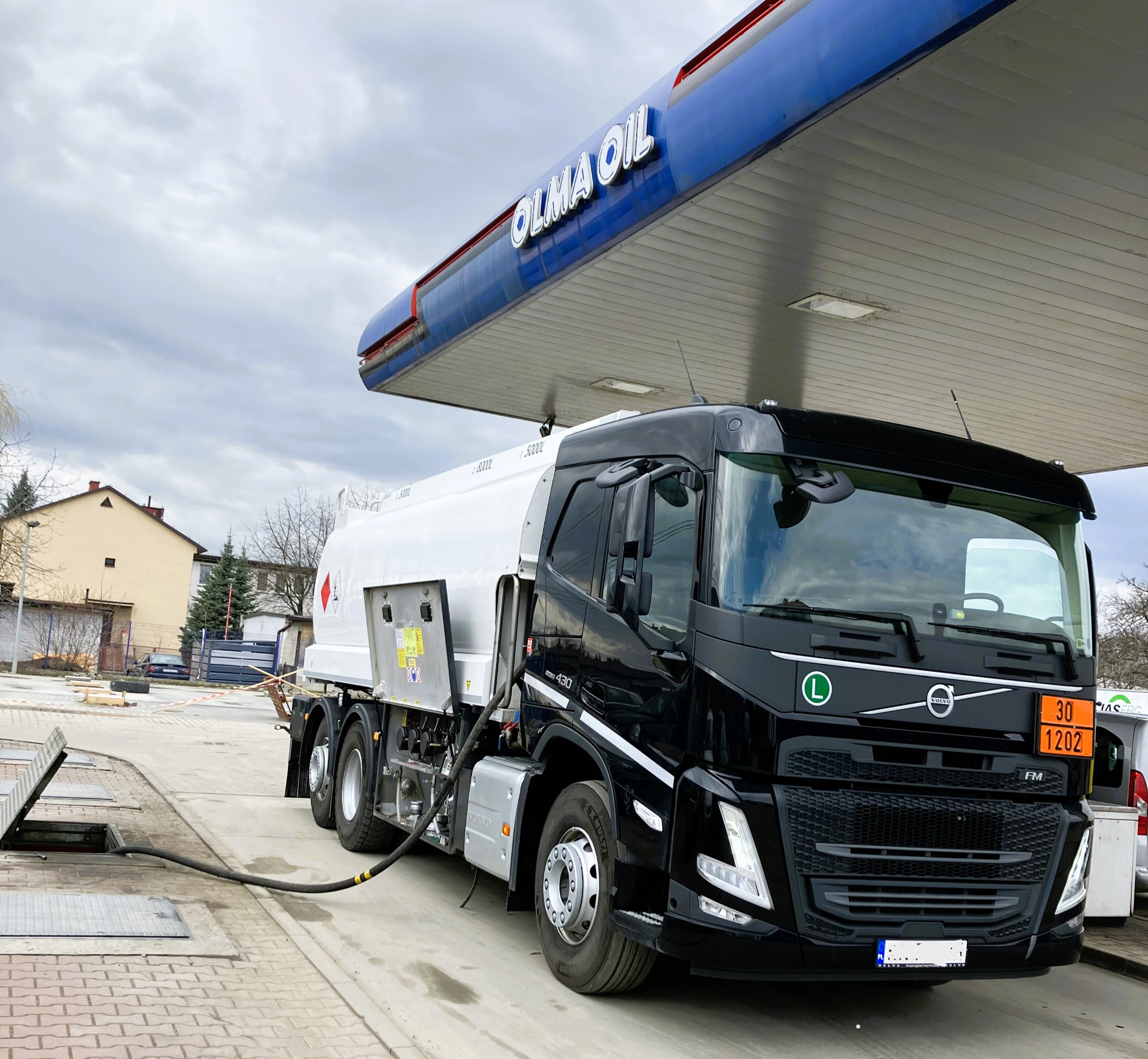 Czarna cysterna Volvo pod dystrybutorem stacji paliw Olma Oil, z podłączonym wężem do podziemnego zbiornika.
