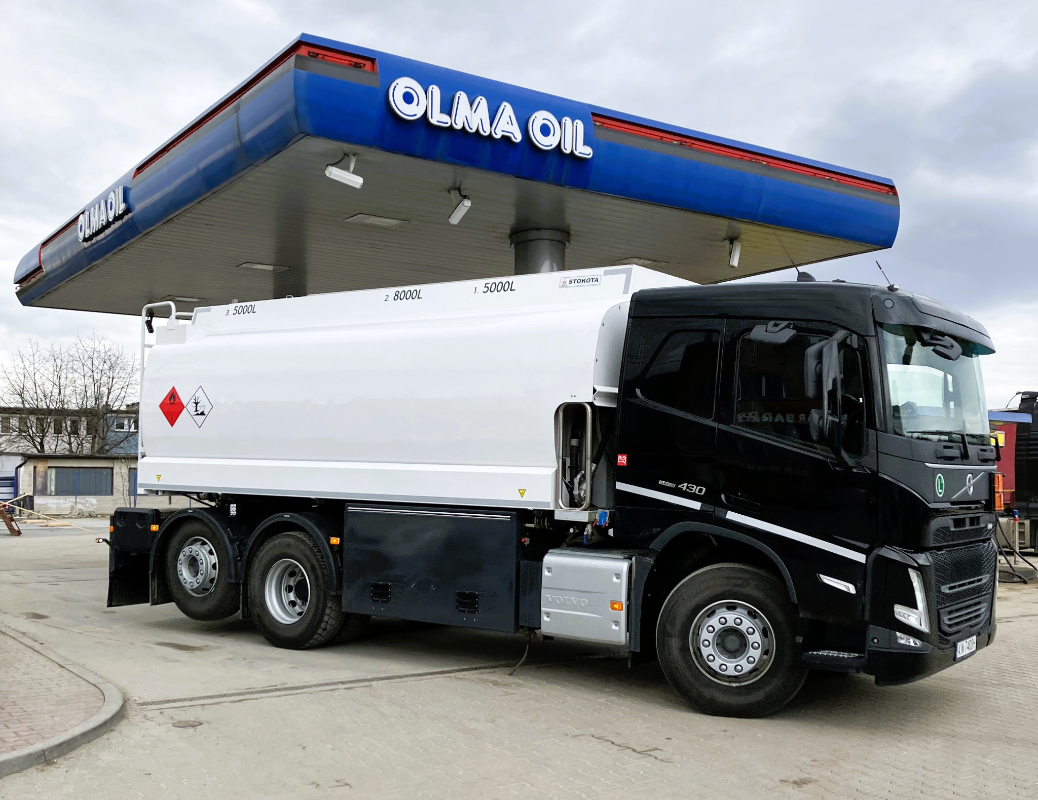 Ciężarówka cysterna marki Volvo z białą cysterną i czarną kabiną, stojąca pod zadaszeniem stacji paliw Olma Oil. Na cysternie widoczne oznaczenia pojemności i symbole ostrzegawcze.