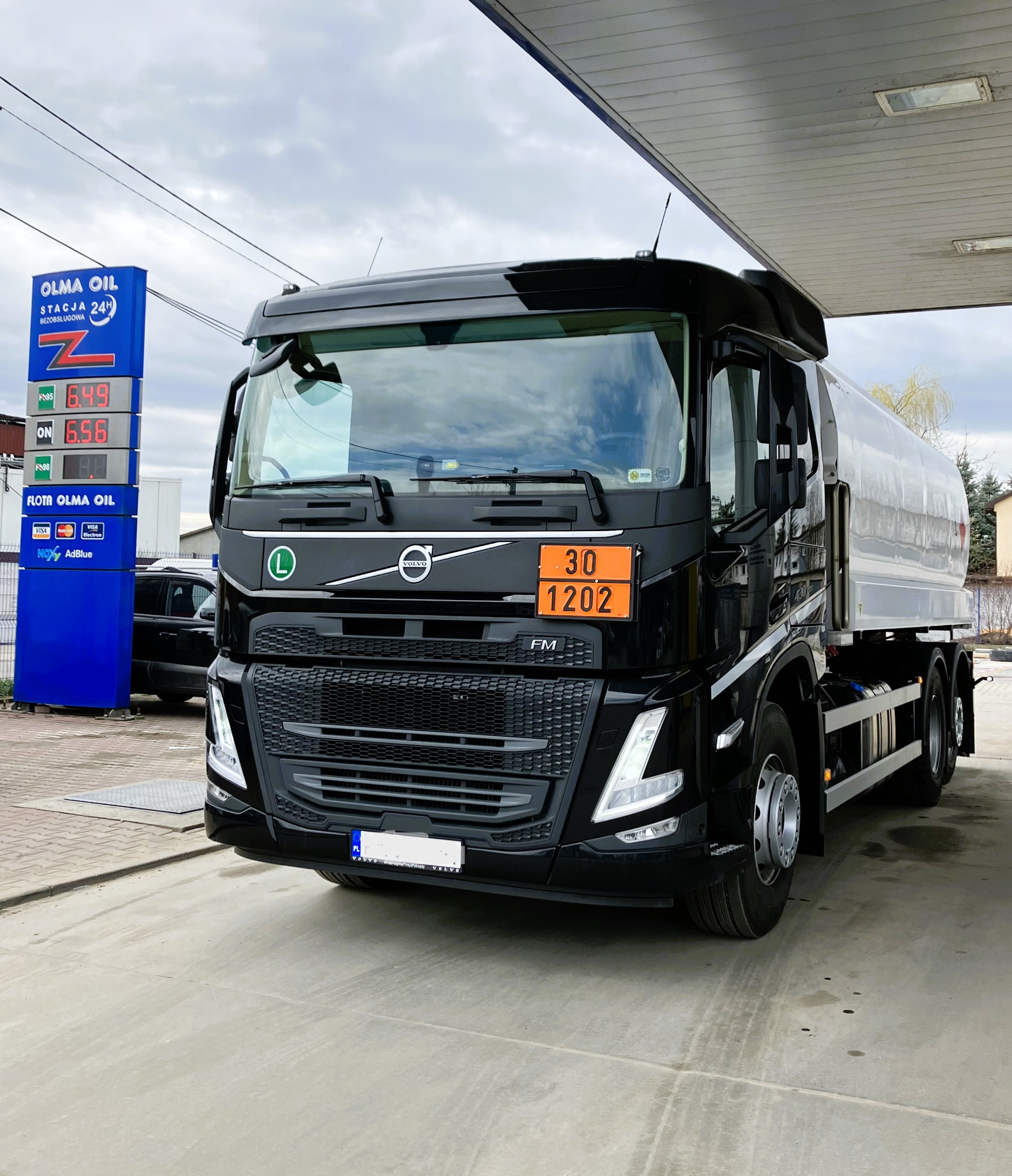 Czarna cysterna Volvo FM z tablicą ostrzegawczą 30/1202 stojąca na stacji paliw Olma Oil, widoczne ceny paliw na pylonie stacji.