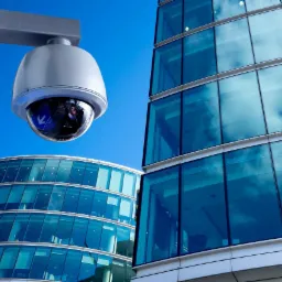 Kopułkowa kamera monitoringu na tle nowoczesnych biurowców z lustrzanymi szybami odbijającymi błękitne niebo.