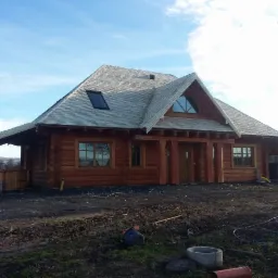 Gotowy dom z bala modrzewiowego o przekroju 300x300mm z szarym dachem gontowym i oknem dachowym, widok z zewnątrz na tle błękitnego nieba.