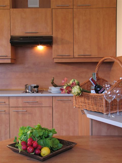 Jasna kuchnia z drewnianymi szafkami, na blacie koszyk piknikowy z butelkami wina i kieliszkami, obok talerze i garnek, na pierwszym planie warzywa i cytryna na drewnianej tacy.