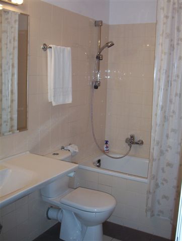 Standardowa łazienka z wanną, toaletą i umywalką, ręcznik wisi na wieszaku, a na wannie stoi butelka płynu do kąpieli.