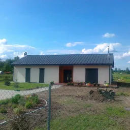 Parterowy dom z ciemnym dachem z blachy, jasną elewacją i drewnianymi elementami. Widok zza ogrodzenia na ogród w słoneczny dzień.