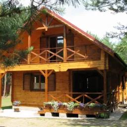 Dwukondygnacyjny dom z bali, z czerwonym dachem, otoczony zielenią. Balkon z drewnianą balustradą, okiennice i kwiaty w donicach dodają uroku.