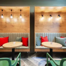 Dwa drewniane boksy z tapicerowanymi siedziskami i oświetleniem, uzupełnione okrągłymi stolikami i zielonymi krzesłami, tworzące przytulną przestrzeń kawiarnianą.