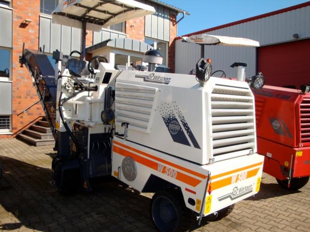 Frezarka do asfaltu Wirtgen W500, biała z pomarańczowymi pasami, zaparkowana przed budynkiem, widoczna z boku, z częściowo wysuniętym ramieniem frezującym i daszkiem przeciwsłonecznym.