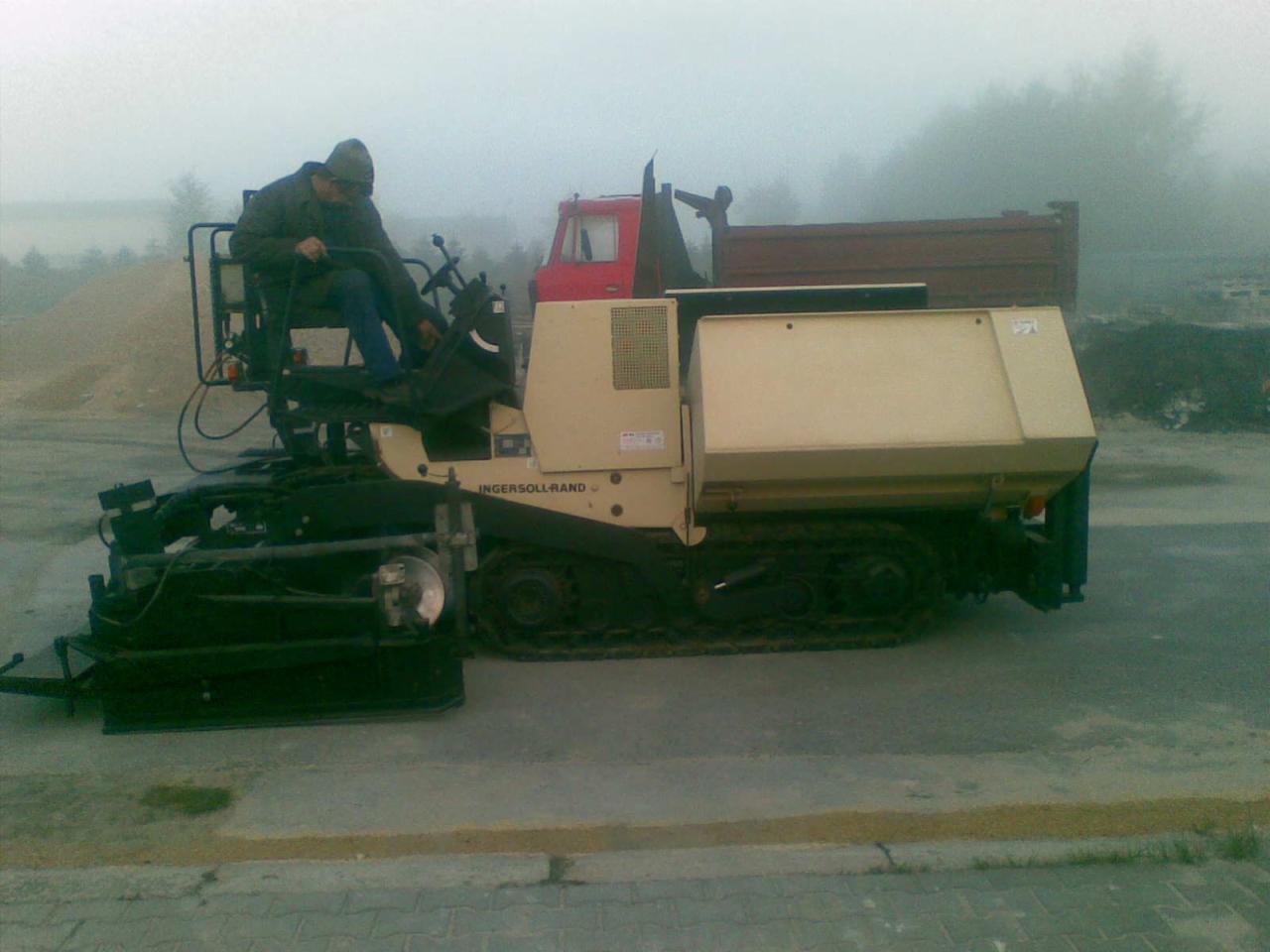 Operator siedzi w maszynie ABG TITAN 111 na gąsienicach, prawdopodobnie rozkładającej asfalt, w tle lekka mgła.