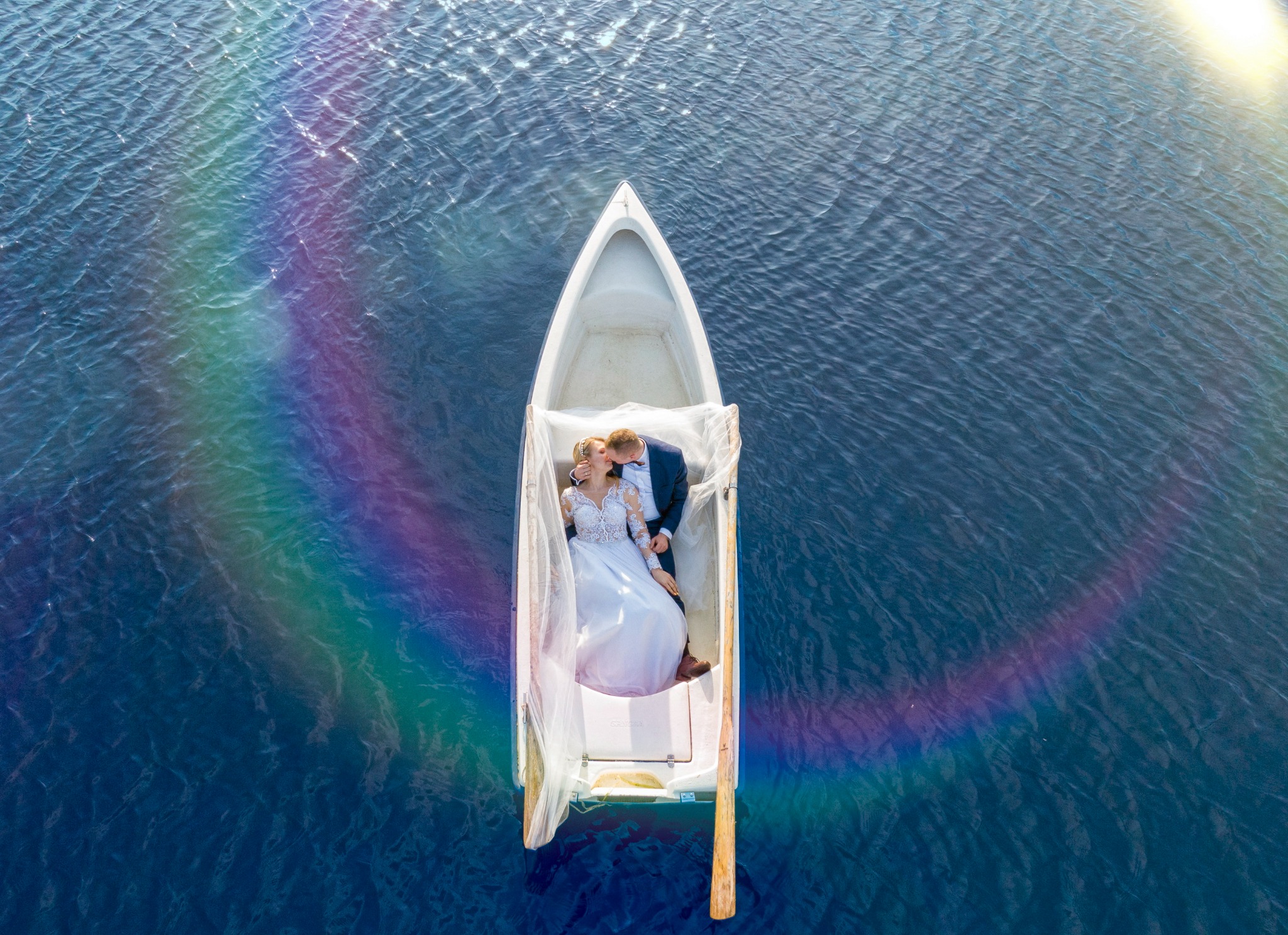 Para młoda w łódce na wodzie, ujęcie z góry, w tle tęcza i odbicia słońca.