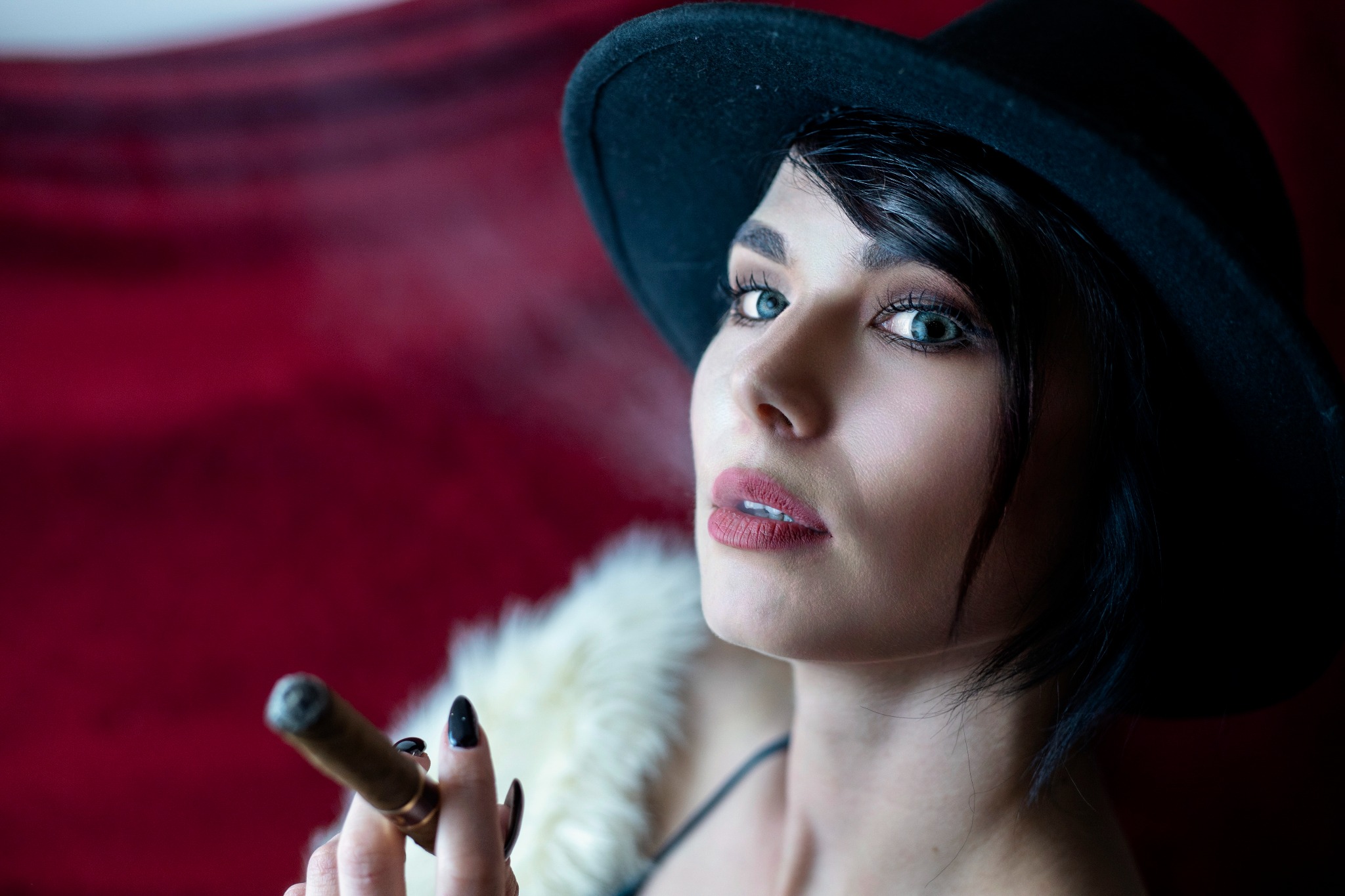 Portret kobiety w czarnym kapeluszu, futrzanym szalu i z cygarem na tle czerwonej tkaniny, z wyraźnym makijażem i niebieskimi oczami.