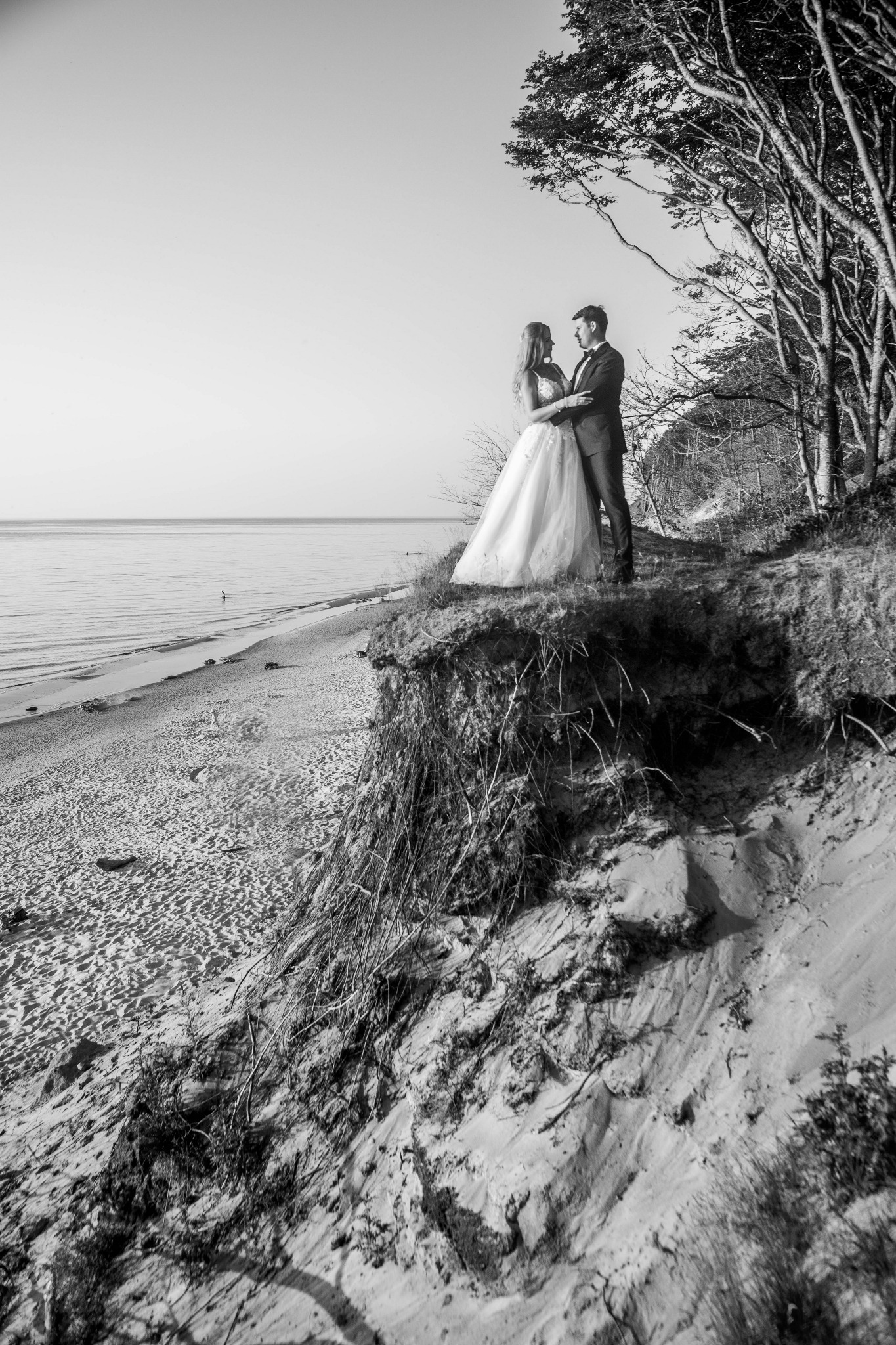 Czarno-białe zdjęcie pary młodej stojącej na klifie z widokiem na morze, panna młoda w sukni z tiulu, pan młody w garniturze, drzewa w tle.