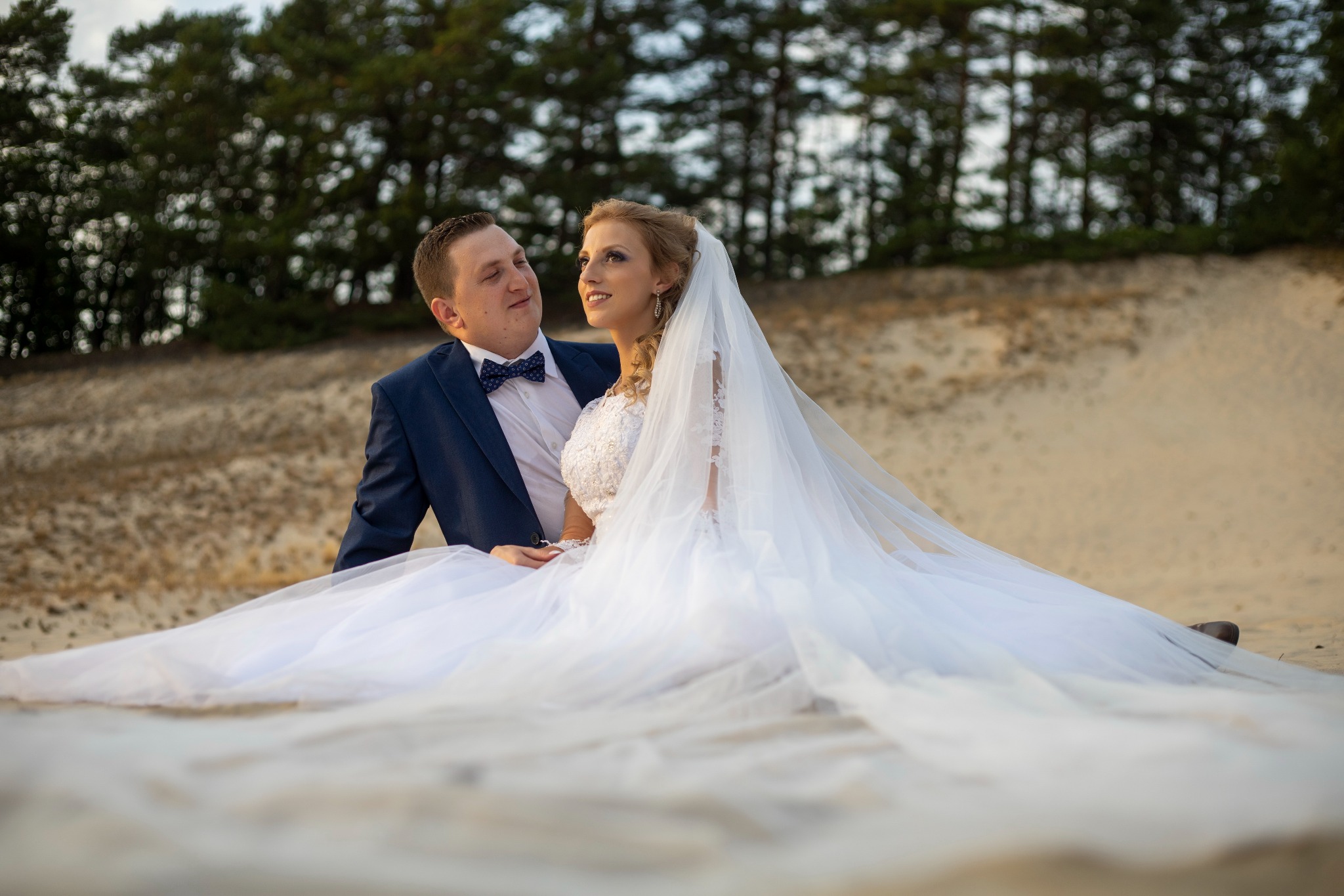 Para młoda siedząca na piasku, on w granatowym garniturze z muszką, ona w białej sukni ślubnej z welonem, spoglądają na siebie z uśmiechem, w tle las