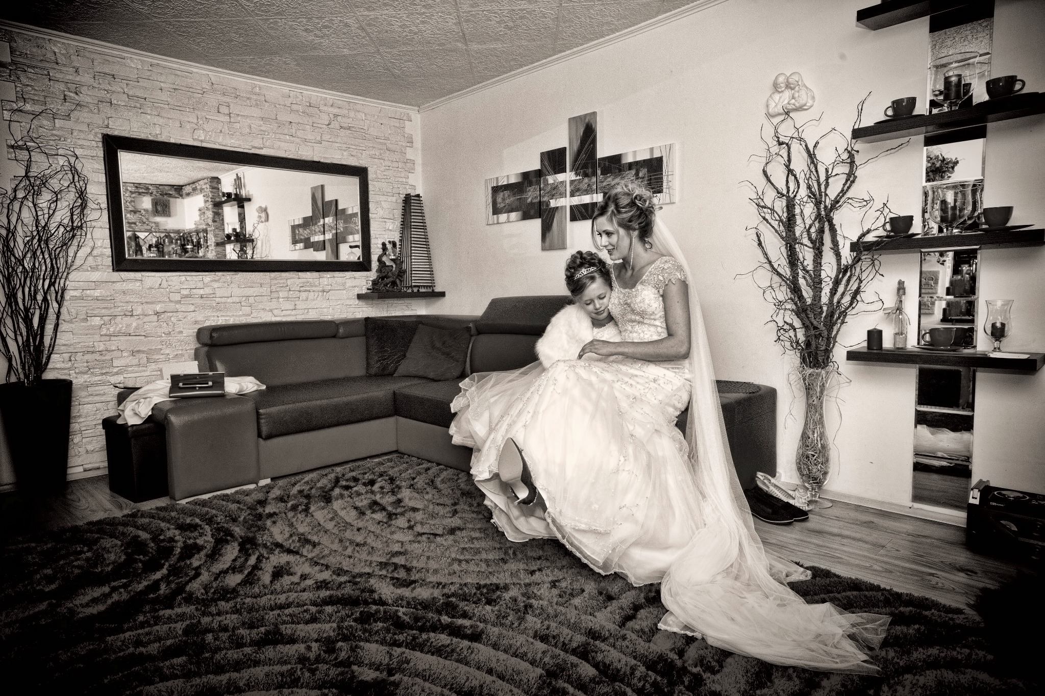 Czarno-białe zdjęcie panny młodej w sukni ślubnej z welonem, siedzącej na kanapie z małą dziewczynką w futerku, w tle ściana z cegły i dekoracje.