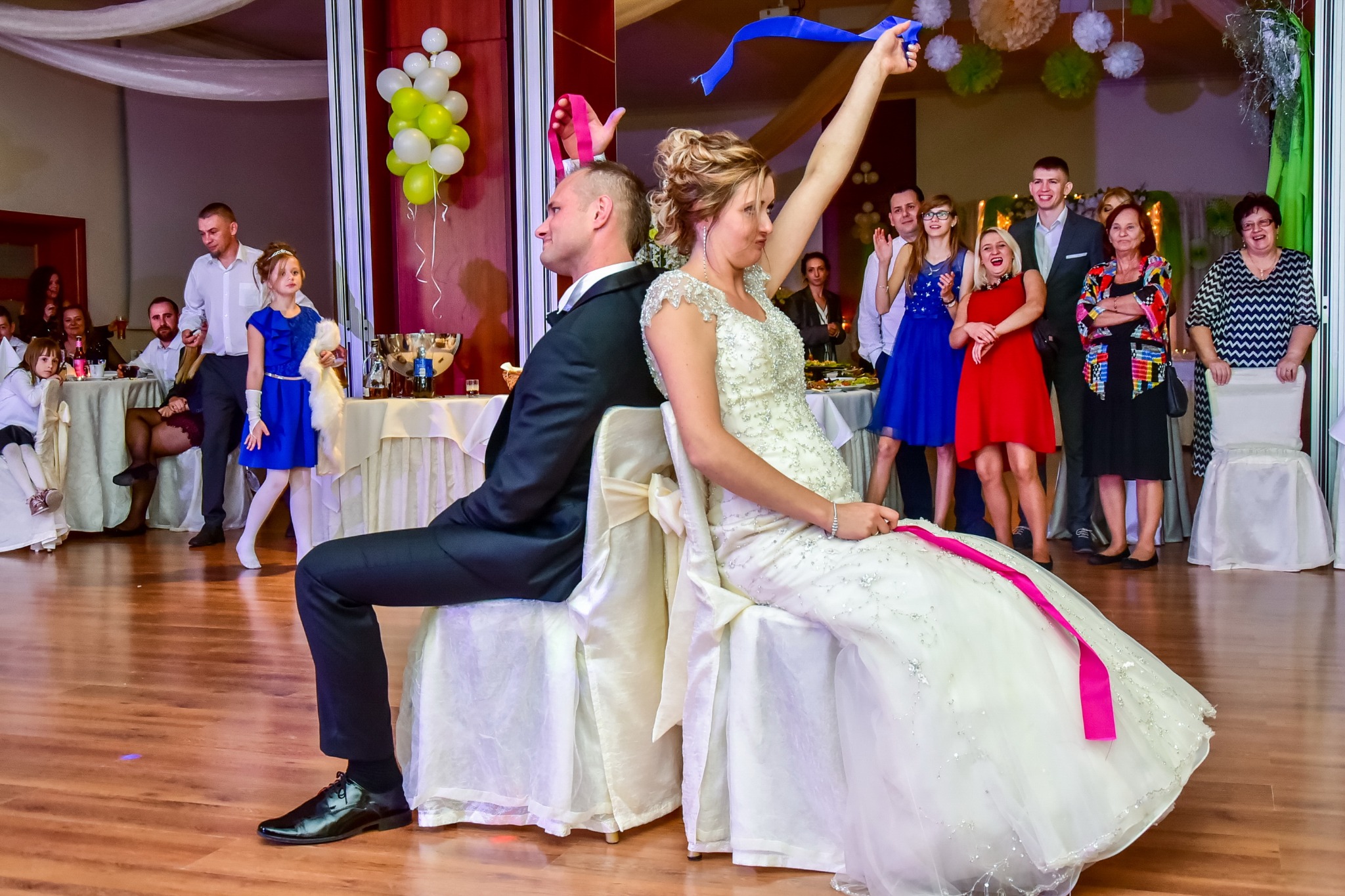 Para młoda siedzi tyłem do siebie na krzesłach podczas oczepin, trzymają w górze wstążki, a w tle goście weselni.