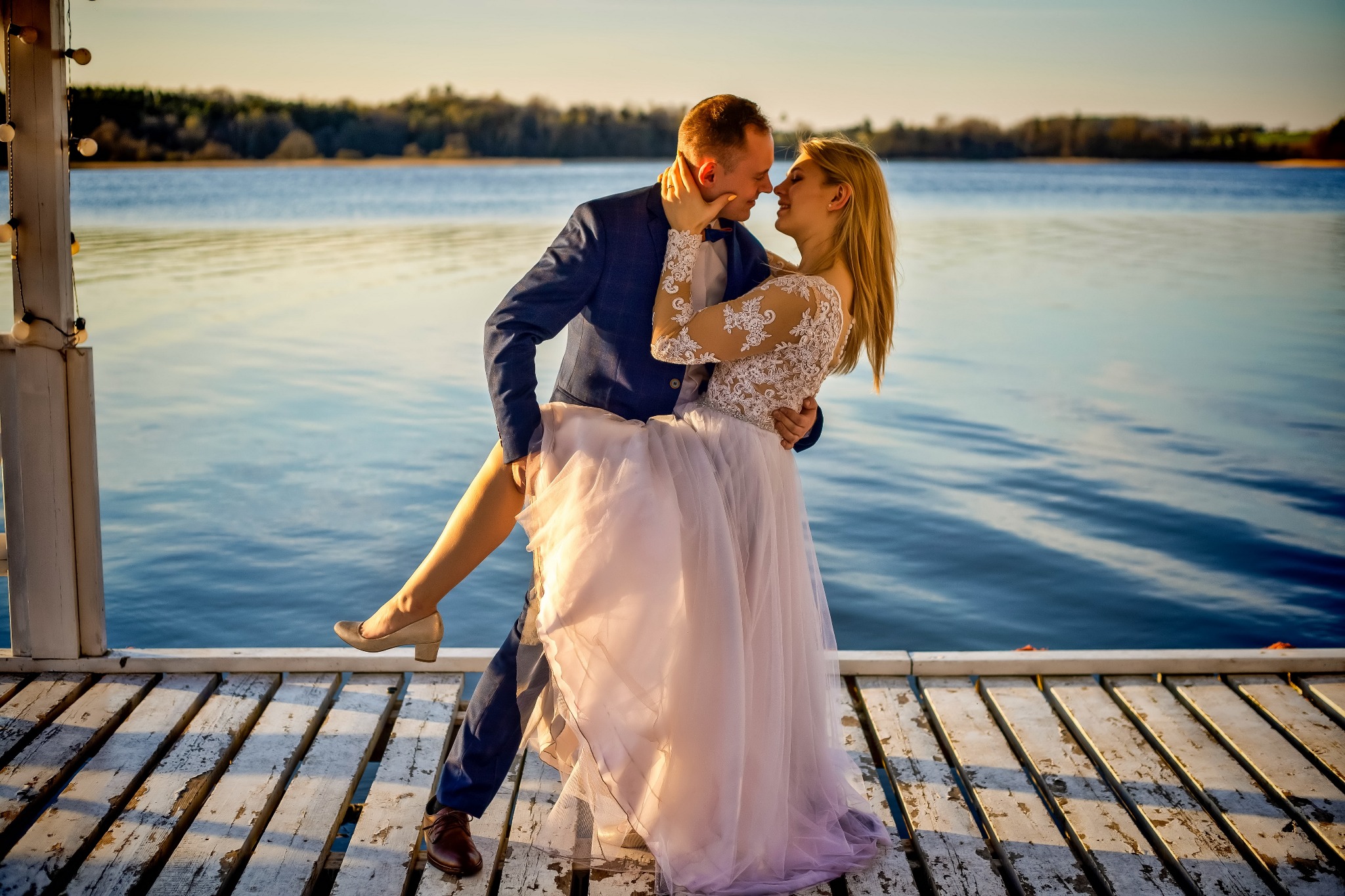 Para młoda, mężczyzna trzyma pannę młodą w objęciach na drewnianym pomoście nad jeziorem, w tle drzewa i zachodzące słońce.