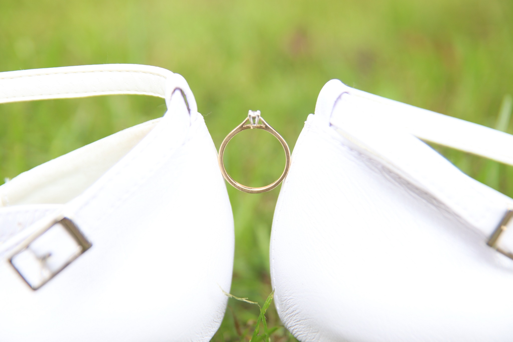 Złoty pierścionek zaręczynowy z brylantem umieszczony pomiędzy dwoma białymi bucikami dziecięcymi na tle zielonej trawy.