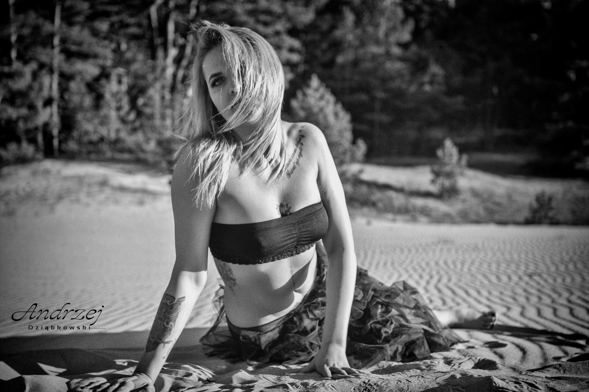 Czarno-białe zdjęcie kobiety leżącej na piasku w bieliźnie i spódnicy, z tatuażami na ciele, włosy opadają na twarz, w tle las.