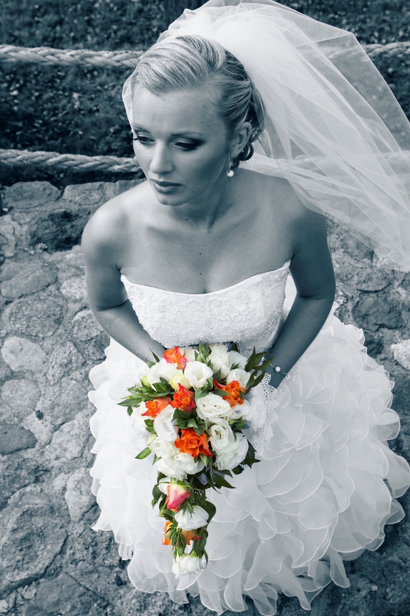 Panna młoda w białej sukni trzyma bukiet białych i pomarańczowych róż, patrząc w dół na tle kamiennych schodów i rozwianego welonu.