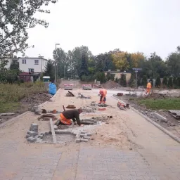 Trzech robotników w pomarańczowych ubraniach układa szarą kostkę brukową na nowej drodze, widoczne narzędzia, palety z kostką, drzewa i budynki w tle.