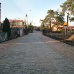 Ulica wyłożona szarą kostką brukową w kształcie litery I, z progami zwalniającymi i znakami drogowymi, otoczona zielenią i budynkami w tle pod błękitnym niebem.