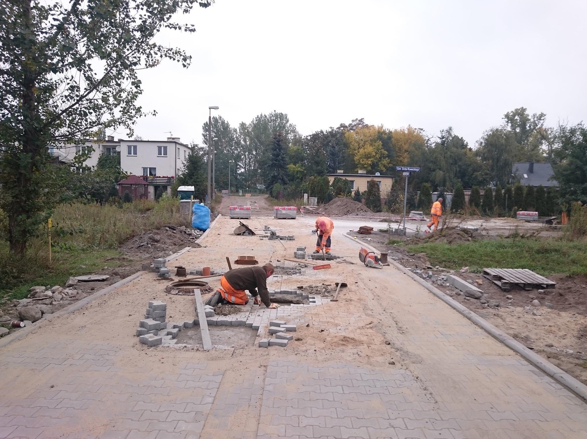Trzech robotników w pomarańczowych ubraniach układa szarą kostkę brukową na nowej drodze, widoczne narzędzia, palety z kostką, drzewa i budynki w tle.