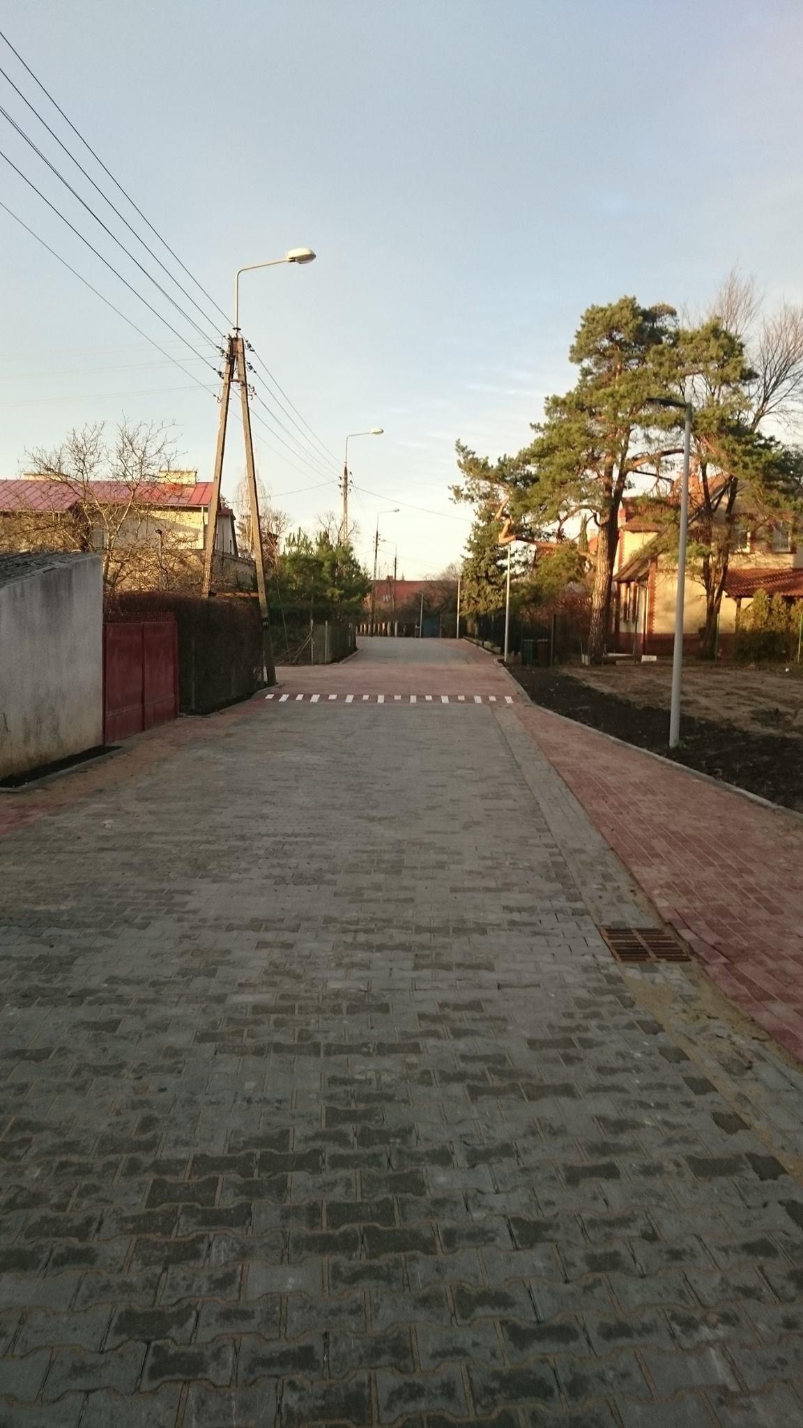 Ulica wyłożona szarą kostką brukową z białymi pasami przejścia dla pieszych, obrzeża z czerwonej kostki, słupy oświetleniowe i drzewa w tle.