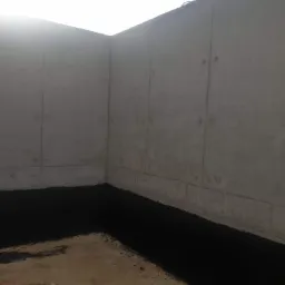 Surowe, betonowe ściany fundamentowe z widoczną izolacją przeciwwilgociową w postaci czarnej powłoki bitumicznej, ziemia na dnie wykopu.