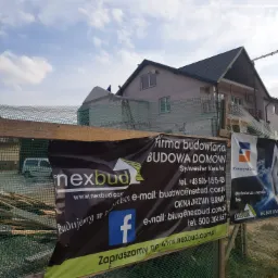 Dom w trakcie budowy, widoczni robotnicy na dachu, na pierwszym planie siatka zabezpieczająca i banery reklamowe firmy budowlanej Nexbud z danymi kontaktowymi i hasłem 'Budujemy w Polsce'.