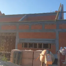 Wznoszenie ścian domu z czerwonej cegły, widoczne betonowe wzmocnienia, drewniane elementy konstrukcyjne, palety cegieł oraz betoniarka na placu budowy w słoneczny dzień.