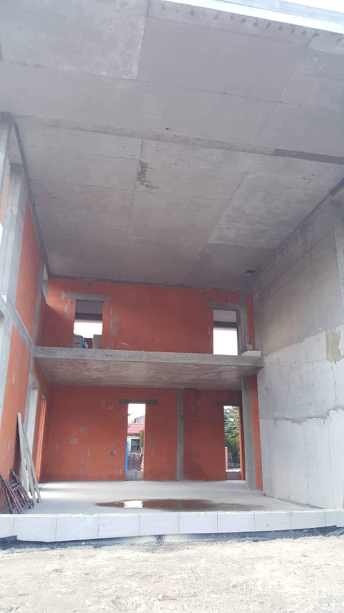 Wnętrze surowego, dwukondygnacyjnego domu w trakcie budowy: ściany z czerwonej cegły, betonowe stropy, widoczne otwory okienne i drzwiowe, na podłodze widoczne kałuże, w tle zieleń i zabudowania.