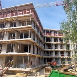 Eco Sense Sp. z o.o. - Wznoszenie wielopiętrowego budynku z betonu komórkowego w otoczeniu zieleni i z widocznym żurawiem budowlanym. Na pierwszym planie pracownicy w kaskach.