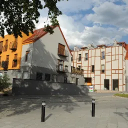 Kompleks budynków o różnorodnej architekturze: pomarańczowa elewacja z balkonami, szara bryła z czerwonym dachem oraz budynek w stylu muru pruskiego z brązowymi belkami.