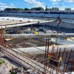 Budowa fundament&oacute;w z rurami ochronnymi i zbrojeniami; widoczni pracownicy i dźwig. Etap prac ziemnych i konstrukcyjnych w otoczeniu miejskim.