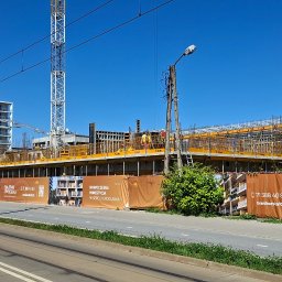 Eco Sense Sp. z o.o. - Budowa apartamentowca w słoneczny dzień. Widoczne rusztowania, dźwigi i ogrodzenie z reklamami inwestycji. Ujęcie z poziomu ulicy, z torami tramwajowymi.